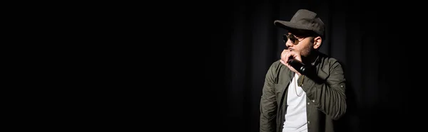 Chanteur hip hop du Moyen-Orient en casquette et lunettes de soleil chantant dans le microphone sur noir, bannière — Photo de stock