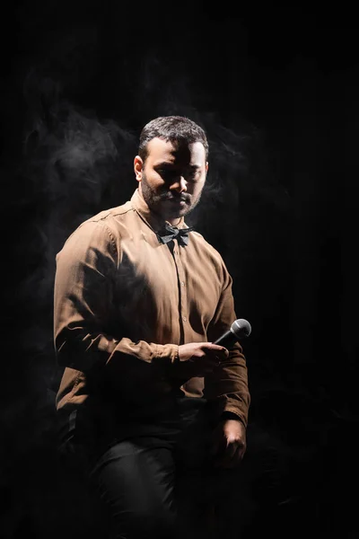 Middle east actor in shirt and bow tie sitting with microphone on black with smoke — Photo de stock