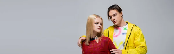 Young man in windbreaker hugging girlfriend and looking at camera isolated on grey, banner - foto de stock