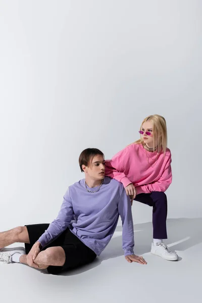 Jolie jeune modèle en lunettes de soleil posant près du petit ami assis sur fond gris — Photo de stock