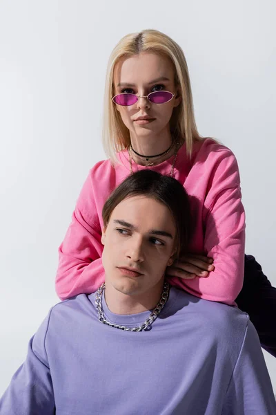 Young blonde model in pink sunglasses looking at camera near boyfriend isolated on grey - foto de stock