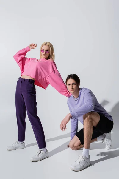 Young woman in pink sweatshirt and sunglasses looking at camera near boyfriend on grey background — Stock Photo