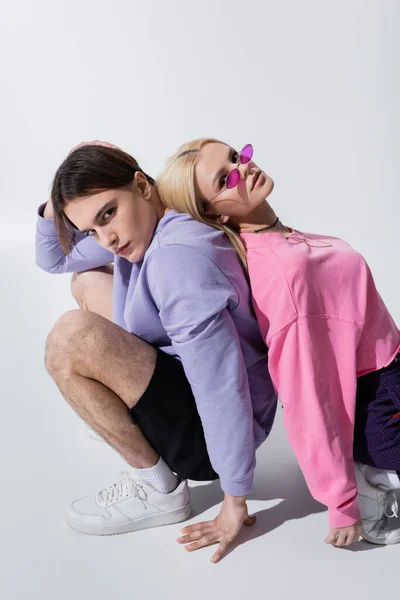 Stilvolles junges Paar blickt in die Kamera, während es Rücken an Rücken auf grauem Hintergrund sitzt — Stockfoto