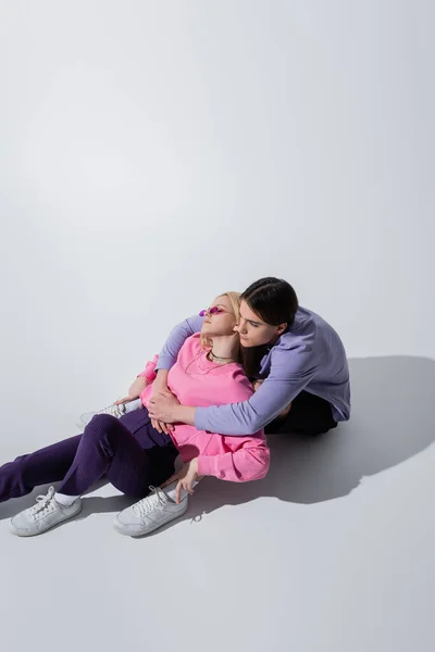Brunette man hugging stylish girlfriend while sitting on grey background — Photo de stock