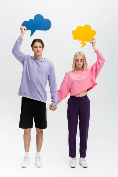 Full length of stylish couple with thought bubbles holding hands on grey background - foto de stock