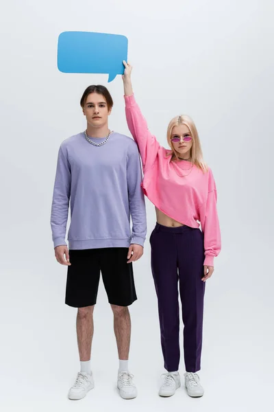 Full length of woman in sunglasses holding speech bubble above boyfriend on grey background - foto de stock