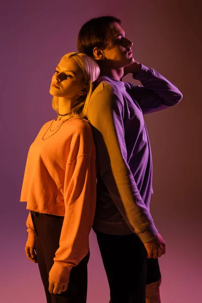 Stylish couple standing back to back on purple background with lighting — Stock Photo