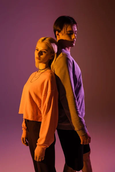 Young couple standing back to back on purple background with lighting — Foto stock