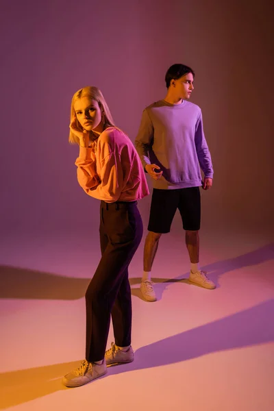 Stylish woman posing near boyfriend in sweatshirt on purple background with lighting — Foto stock