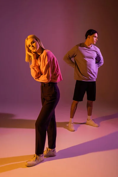 Junge blonde Frau schaut in die Kamera in der Nähe brünette Freund auf lila Hintergrund mit Beleuchtung — Stockfoto