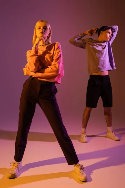 Trendy young woman looking at camera near boyfriend on purple background with lighting — Foto stock