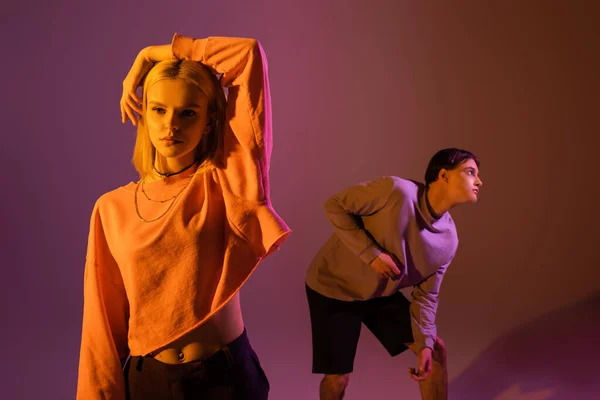 Blonde woman touching hair near boyfriend on purple background with lighting — Photo de stock