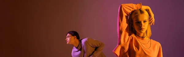 Stylish woman touching blonde hair near blurred boyfriend on purple background with lighting, banner - foto de stock
