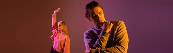 Стильний молодий чоловік позує біля розмитої дівчини на фіолетовому фоні з освітленням, банер — стокове фото