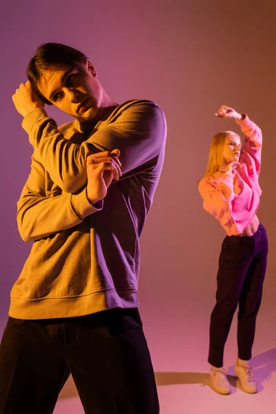 Stylish young man in sweatshirt posing near blurred girlfriend on purple background with lighting — Foto stock