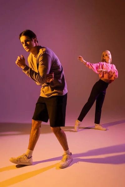 Full length of stylish couple in sweatshirts and sneakers on purple background with lighting — Stockfoto