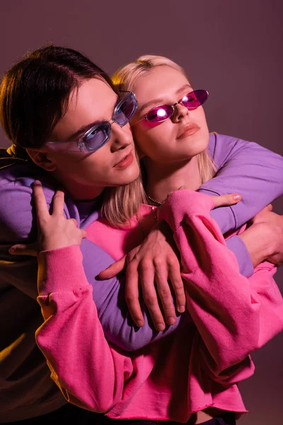 Stilvoller Mann mit Sonnenbrille umarmt Freundin isoliert auf lila mit Beleuchtung — Stockfoto