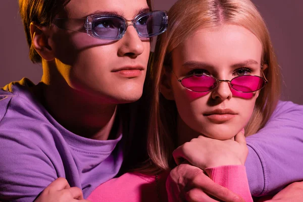 Portrait of man embracing girlfriend in sweatshirt and sunglasses isolated on purple with lighting - foto de stock