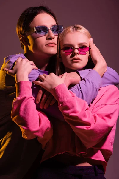 Stylish couple in sunglasses hugging and looking at camera isolated on purple with lighting — Photo de stock
