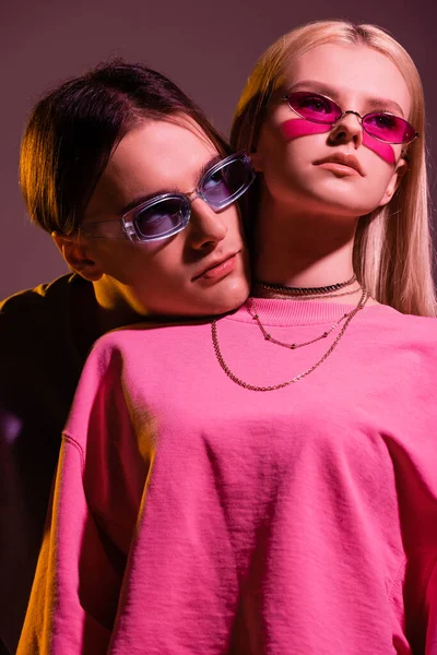 Young couple in sunglasses and sweatshirts looking away isolated on purple with lighting — Foto stock