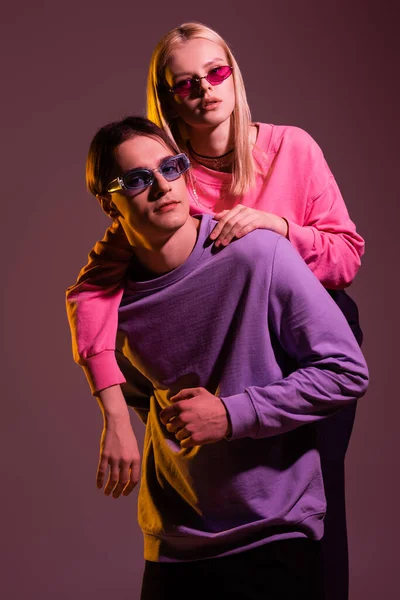 Mulher na moda em óculos de sol abraçando namorado em camisola isolada em roxo com iluminação — Fotografia de Stock