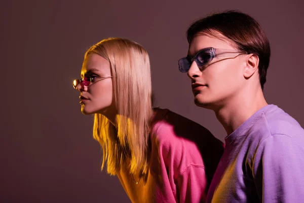 Couple élégant en lunettes de soleil regardant loin isolé sur violet avec éclairage — Photo de stock