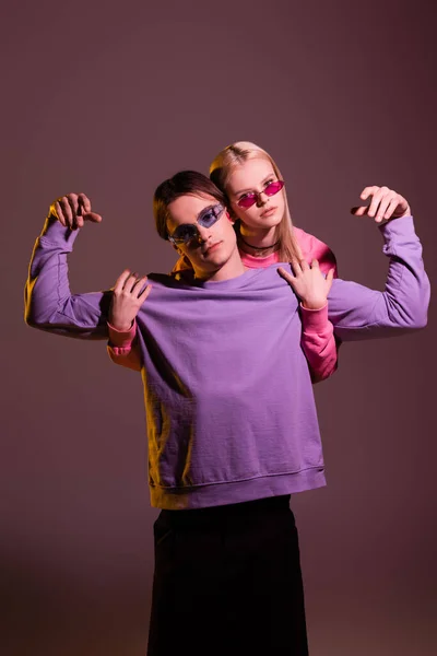 Blonde woman in sunglasses hugging stylish boyfriend isolated on purple with lighting — Stock Photo