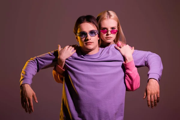 Blonde woman in pink sunglasses hugging boyfriend isolated on purple with lighting — Stockfoto