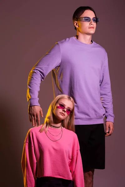Giovane uomo in occhiali da sole e felpa posa vicino fidanzata su sfondo viola con illuminazione — Foto stock