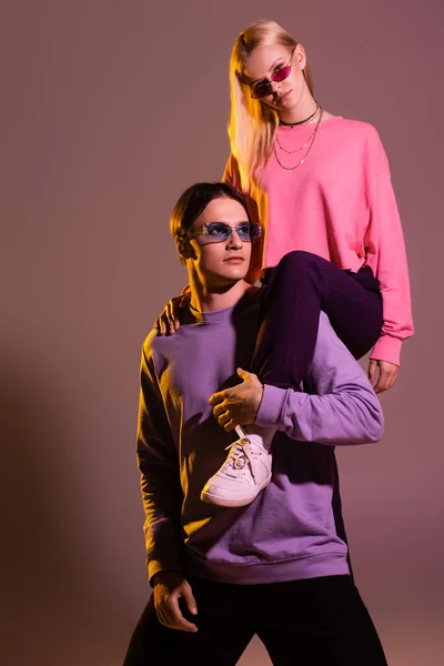 Stilvolle Frau mit Sonnenbrille posiert mit Freund auf lila Hintergrund mit Beleuchtung — Stockfoto