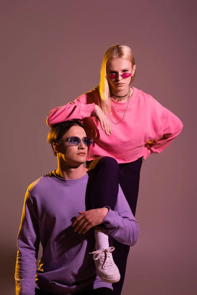 Pretty blonde woman looking at camera near boyfriend in sunglasses on purple background with lighting — Fotografia de Stock