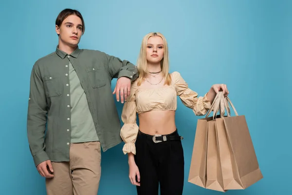 Mujer joven sosteniendo bolsas de compras cerca de novio elegante sobre fondo azul - foto de stock