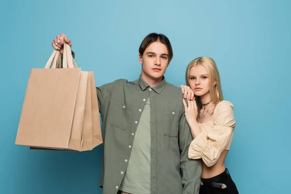 Hübsche blonde Frau umarmt Freund mit Einkaufstaschen auf blauem Hintergrund — Stockfoto