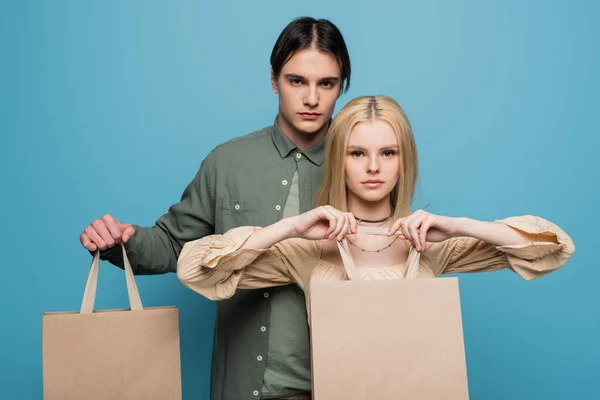 Morena hombre mirando a la cámara cerca de novia con bolsas de compras aisladas en azul - foto de stock