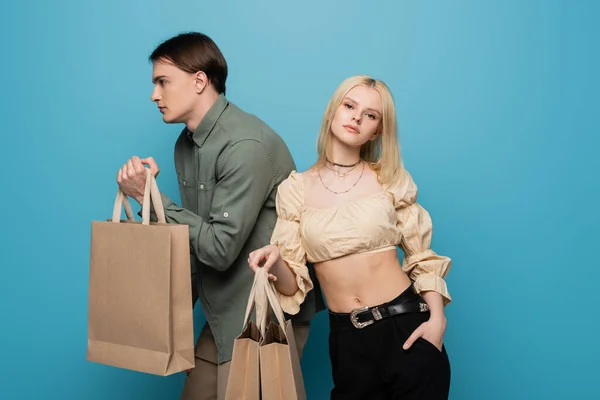 Mujer joven en blusa posando con bolsas de compras cerca de novio sobre fondo azul - foto de stock