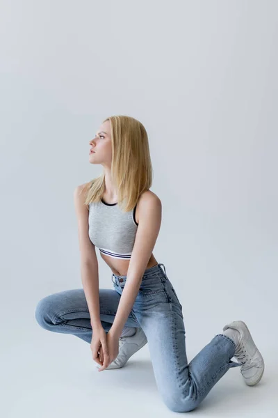 Vue latérale de la femme blonde en haut et jeans regardant loin sur fond gris — Photo de stock