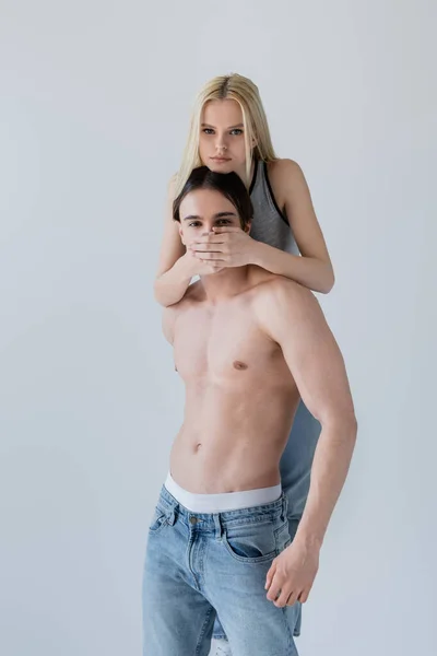 Hübsche blonde Frau bedeckt Mund ihres hemdlosen Freundes in Jeans isoliert auf grau — Stockfoto