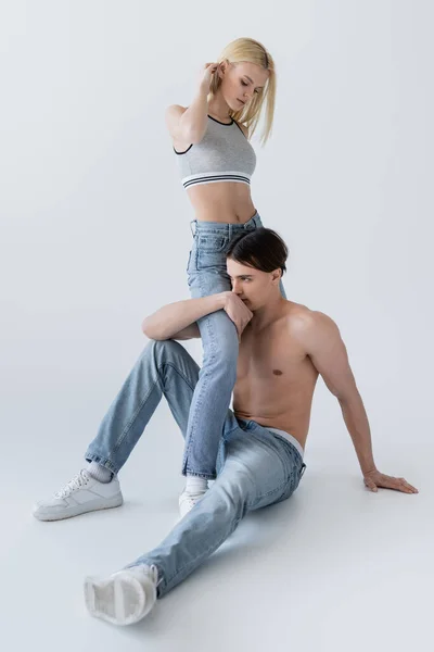 Full length of shirtless man in jeans hugging leg of blonde girlfriend on grey background - foto de stock