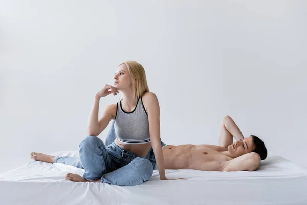 Мускулистый мужчина лежит на кровати рядом с подругой в джинсах и топ изолирован на сером — стоковое фото