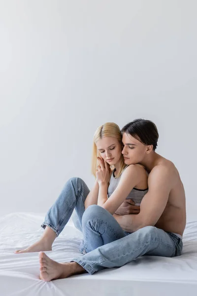 Uomo muscoloso che abbraccia la ragazza bionda in jeans sul letto isolato sul grigio — Foto stock