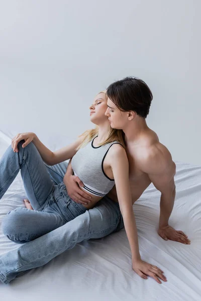 Sexy hombre abrazando novia en la parte superior y pantalones vaqueros en la cama aislado en gris - foto de stock