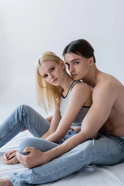 Stylish couple in jeans looking at camera on bed isolated on grey — Stockfoto