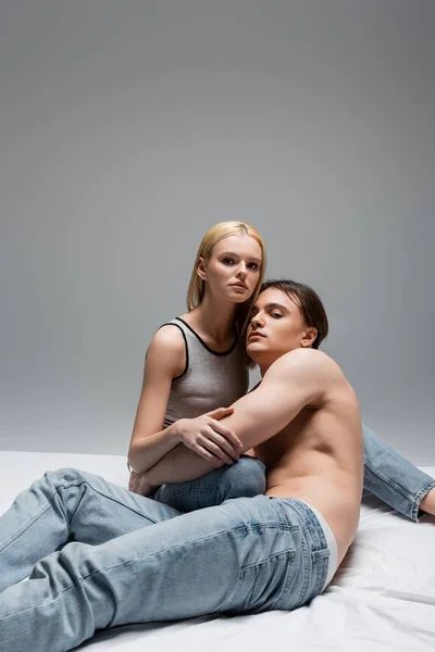 Muskulöser Mann in Jeans umarmt Freundin und schaut in Kamera auf Bett isoliert auf grau — Stockfoto