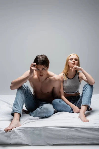 Young couple in jeans posing while sitting on bed on grey background — Fotografia de Stock