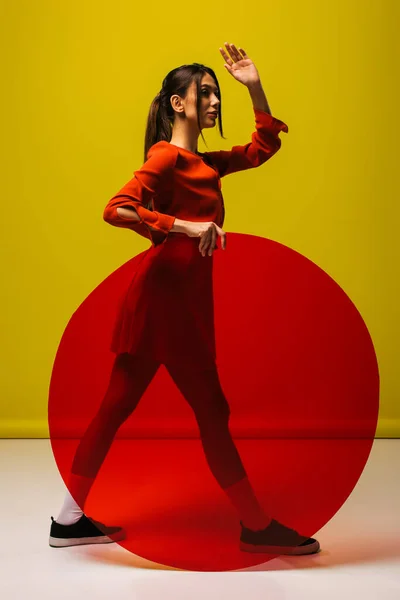 Volle Länge der stilvollen jungen Frau in rotem Kleid, die rundes Glas in Form hält und auf Grün gestikuliert — Stockfoto