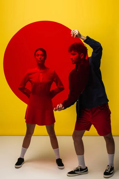 Trendy young man holding round shape glass near woman in dress posing on yellow — стоковое фото