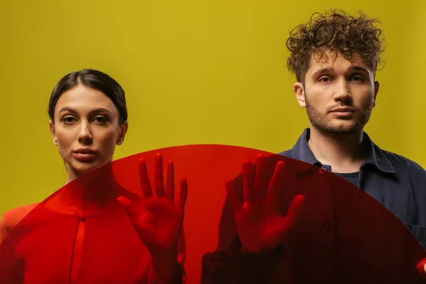 Modelos modernos e jovens posando perto de forma redonda vidro vermelho isolado no verde — Fotografia de Stock