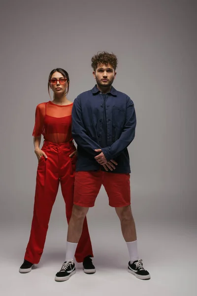 Comprimento total de casal elegante em roupas da moda e óculos de sol posando em cinza — Fotografia de Stock