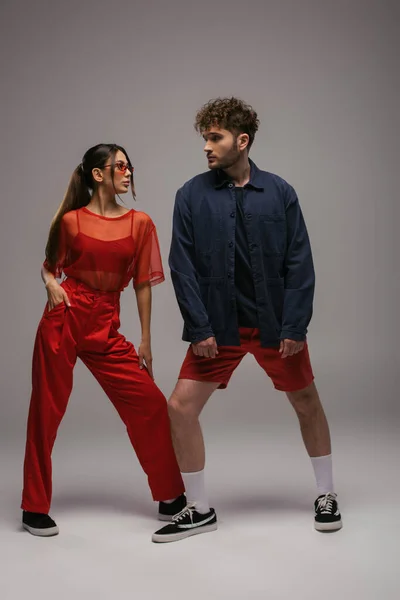 Comprimento total de modelos elegantes em roupas da moda e óculos de sol posando em cinza — Fotografia de Stock