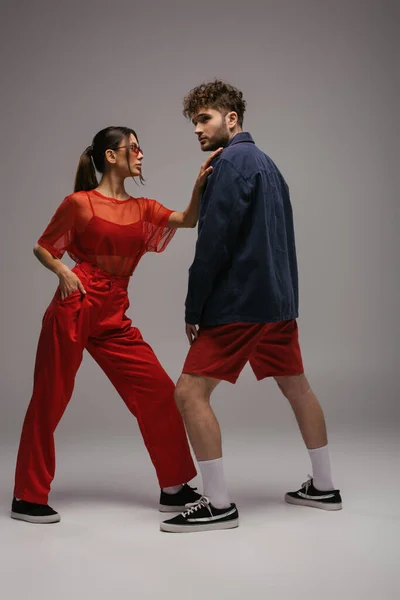 Comprimento total da mulher elegante na roupa da moda e óculos de sol posando perto do homem em cinza — Fotografia de Stock